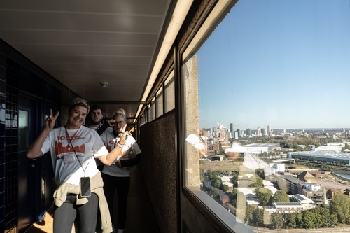 Group of people wearing '10 Towers Challenge' t-shirts walk along a high-rise corridor, with one woman flashing a peace sign, while a city skyline is visible through the wind