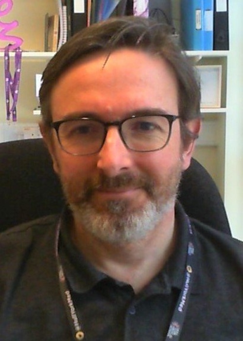 Man with glasses and a beard, wearing a lanyard, seated in an office