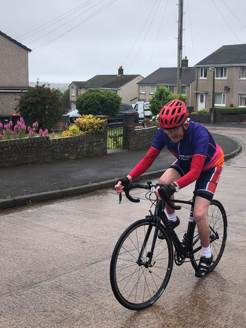 Man wearing a red helmet and an Asthma + Lung UK cycling jersey rides a black road bike on a wet suburban street lined with houses and gardens