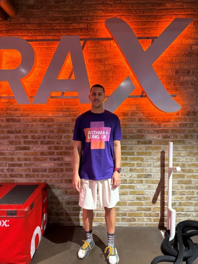 Man wearing an Asthma + Lung UK t-shirt and sportswear stands in front of a brick wall with a large illuminated sign at a gym
