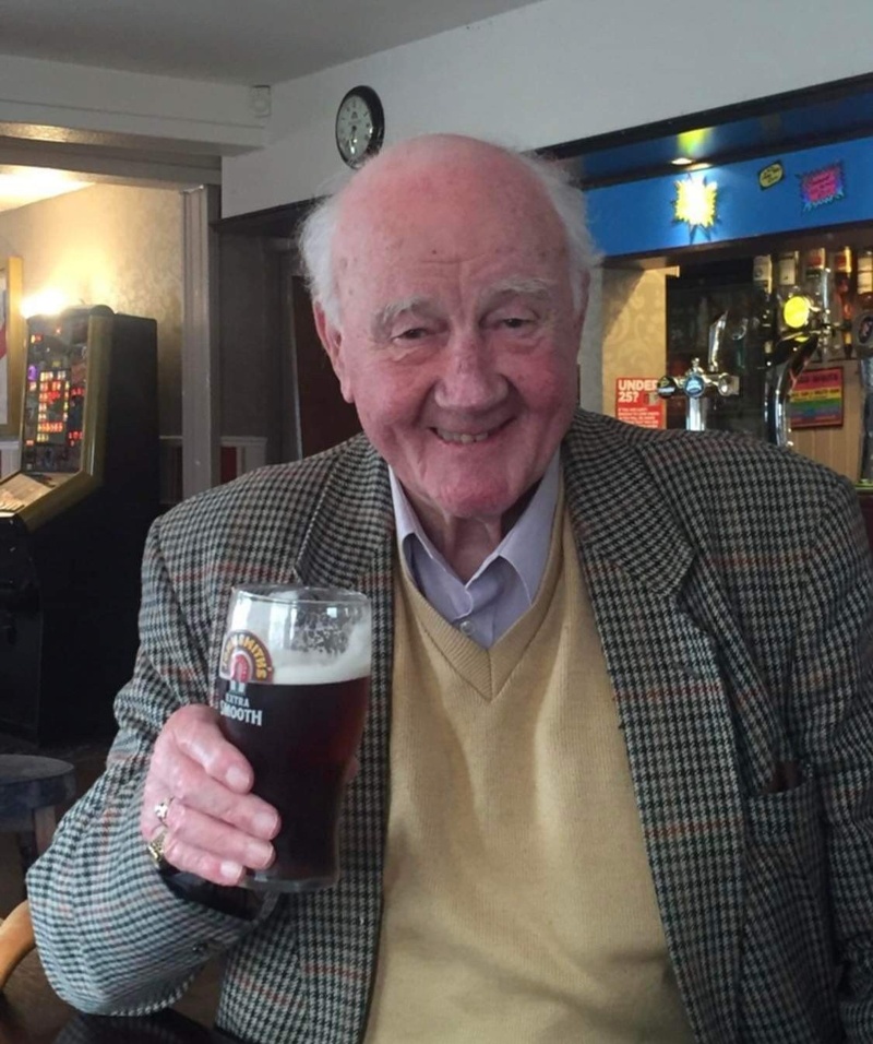 Smiling elderly man in a tweed jacket and sweater holds up a pint of beer while sitting in a pub