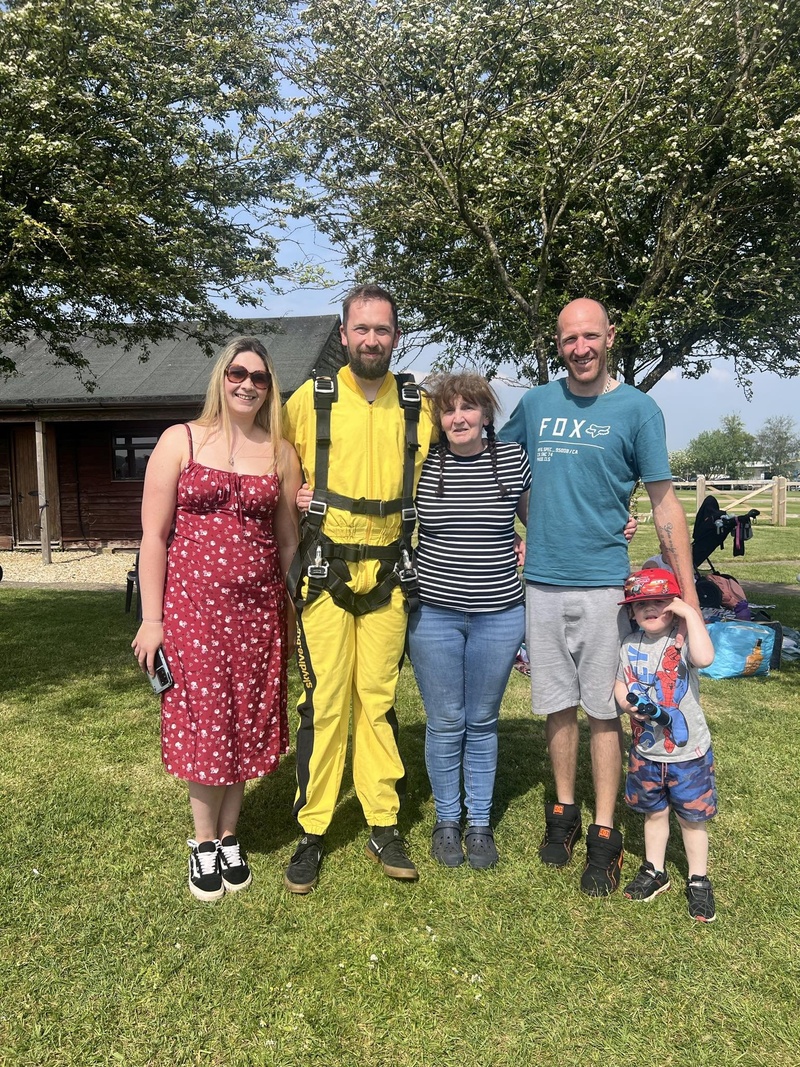ive people stand together outdoors on a sunny day, with one man wearing a yellow skydiving suit, surrounded by family members, including a young child in a Spider-Man cap