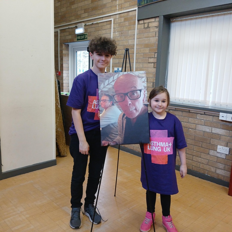 Two children wearing Asthma + Lung UK t-shirts stand beside a large photo of a man with glasses on an easel in a community hall