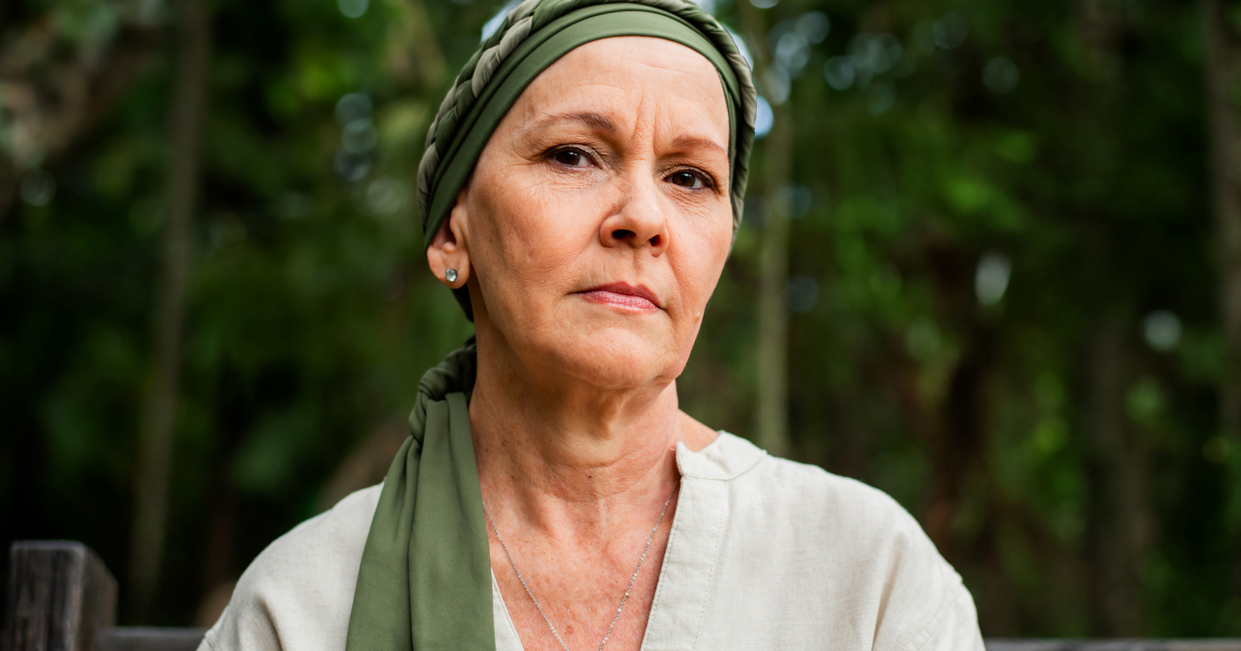 A woman with a headscarf and a serious expression, standing outdoors with a blurred green background