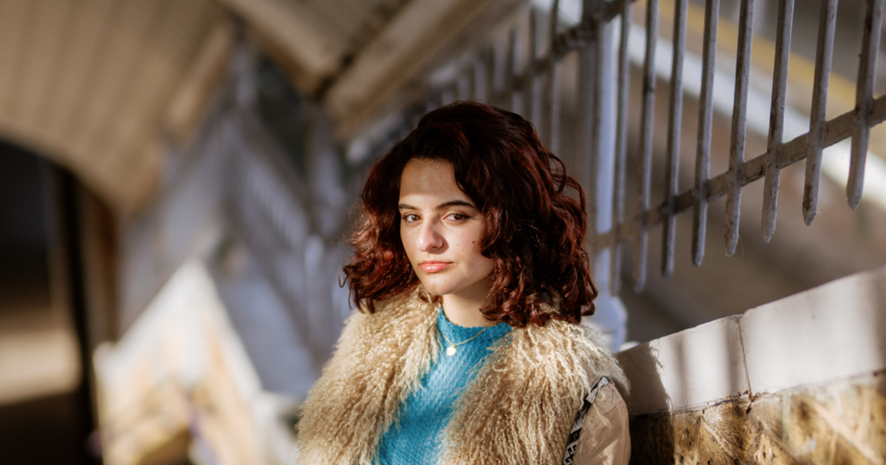 A woman with curly dark hair, in a beige fur-trimmed coat, stands on a staircase, looking thoughtful