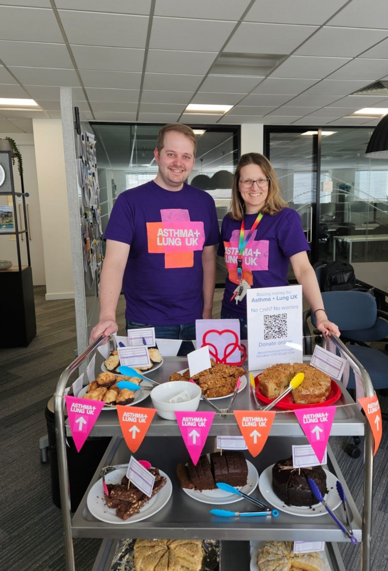 Two smiling people wearing Asthma + Lung UK t-shirts stand behind a cart filled with homemade cakes and treats, raising money for charity in an office setting
