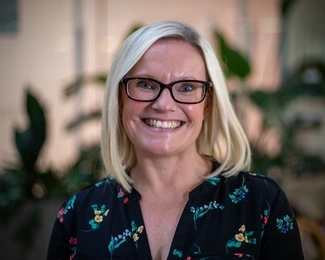 A smiling woman with blonde hair and glasses wearing a floral blouse, with blurred green plants in the background