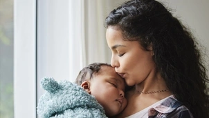 A mother gently kisses her sleeping baby’s forehead while holding them close by a window