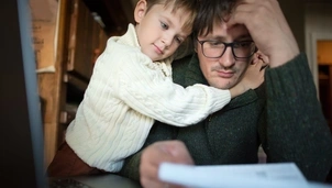 A stressed man reviews documents as a child embraces him from behind