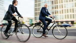 Two men in business attire cycle through a city street with blurred buildings and traffic in the background