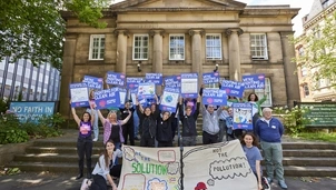 A group of campaigners stand outside a historic building, holding signs that read Fighting for Clean Air and banners promoting clean air solutions