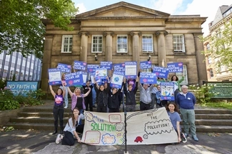 A group of campaigners stand outside a historic building, holding signs that read Fighting for Clean Air and banners promoting clean air solutions