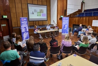 A group of people attending a health conference, listening to speakers at the front of the room with banners promoting the cause