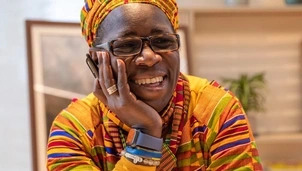 A smiling woman in vibrant, patterned clothing and glasses rests her head on her hand