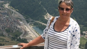 A woman wearing sunglasses and a striped top stands at a scenic viewpoint with mountains, greenery, and a winding road below