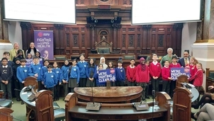 A group of school children in blue and red uniforms stand inside a grand council chamber holding signs that read "We're fighting for clean air," with banners for Asthma + Lung UK in the background