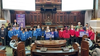 A group of school children in blue and red uniforms stand inside a grand council chamber holding signs that read "We're fighting for clean air," with banners for Asthma + Lung UK in the background