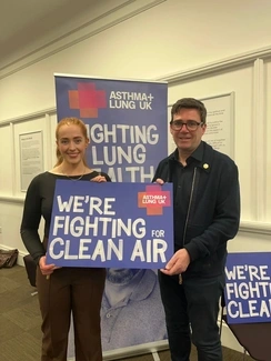 A man and a woman stand indoors holding a sign that reads "We're fighting for clean air," with Asthma + Lung UK banners in the background.