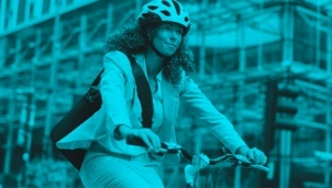 A woman wearing a helmet and a business suit rides a bicycle through an urban area with scaffolding in the background, tinted in blue