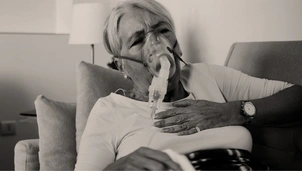 A woman sits on a sofa with one hand on her chest, wearing an oxygen mask, looking distressed