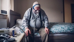 An elderly man sits on the edge of his bed wearing a mask connected to a tube, with equipment and personal items on the bedside table