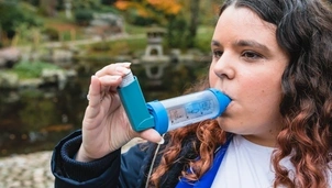 A woman with long, wavy brown hair uses an inhaler with a spacer in an outdoor park setting with autumn trees and a pond in the background