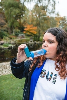 A woman with long, wavy brown hair uses an inhaler with a spacer in an outdoor park setting with autumn trees and a pond in the background