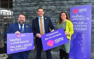 Three people stand in front of a brick wall holding signs supporting lung health, with banners for Asthma + Lung UK Cymru in English and Welsh in the background