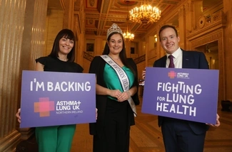 Three people stand in an ornate hall holding signs supporting lung health, with a woman in a sash and tiara at the centre, and banners for Asthma + Lung UK Northern Ireland in the background