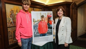 A young man in a pink hoodie stands next to a woman in a white blazer, both smiling beside a large portrait of him displayed on an easel in a wood-panelled room