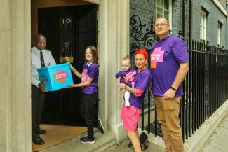 A young girl in a purple Asthma + Lung UK T-shirt hands a blue box to a security officer at the entrance of 10 Downing Street, while three other supporters, including a woman holding a baby, stand nearby