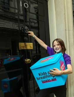 A young girl in a purple Asthma + Lung UK T-shirt holds a blue box with the message Kids are breathing toxic air, while knocking on the black door of 10 Downing Street