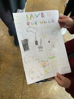 A child holds up a hand-drawn poster titled SAVE OUR WORLD, depicting a split image comparing pollution with a cleaner future