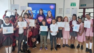A group of schoolchildren and two adults hold up their drawings while standing in front of a screen displaying an Asthma + Lung UK logo