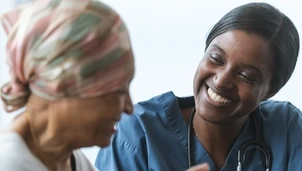 A smiling healthcare professional in blue scrubs and a stethoscope engages warmly with a patient wearing a headscarf