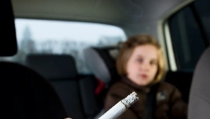 A lit cigarette is held in the foreground inside a car, with a young child in a car seat in the blurred background