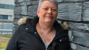 A woman with short grey hair wearing a black winter coat with a fur-trimmed hood, leaning against a textured stone wall