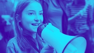 A young girl smiles while speaking into a megaphone at a public gathering, with a blurred crowd in the background