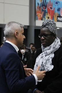 A man in a suit and a woman wearing glasses and a patterned headscarf engage in a serious conversation, with a group of people in the background