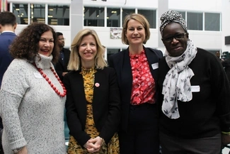 Four women stand together, smiling at the camera in a bright indoor setting with other people in the background