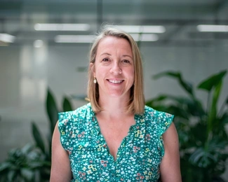 Smiling woman with shoulder-length blonde hair wearing a green floral dress, standing in an office with plants in the background