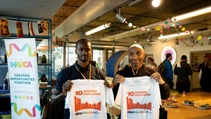 Two men wearing black polo shirts hold up white '10 Towers Challenge' t-shirts in a decorated indoor space with people, food, and a Poplar HARCA banner in the background