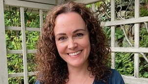 A woman with curly gray hair sits on a couch, looking pensive with her hand resting on her temple