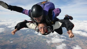 Smiling woman tandem skydives with an instructor above a landscape of fields and clouds, arms outstretched in excitement