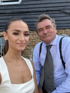 Young woman in a white dress and gold necklace takes a selfie with an older man in a blue shirt and patterned tie, both smiling against a rustic brick and wood building backdrop
