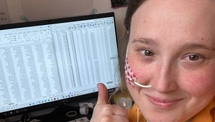 Smiling woman with a nasal tube gives a thumbs-up while working at her desk with spreadsheets open on a computer screen