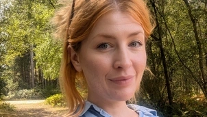 Smiling woman with red hair and sunglasses on her head takes a selfie on a sunlit forest path, wearing a light blue button-up shirt.