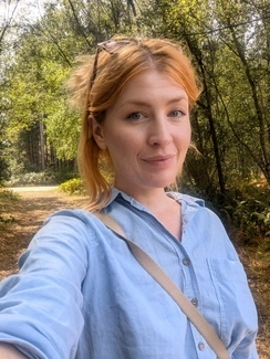 Smiling woman with red hair and sunglasses on her head takes a selfie on a sunlit forest path, wearing a light blue button-up shirt.
