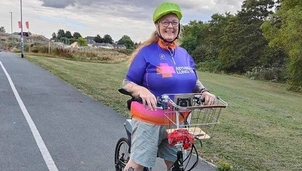 Smiling woman wearing a green helmet and an Asthma + Lung UK cycling jersey stands with her bicycle on a paved path, with a basket attached to the front and a scenic landscape behind her
