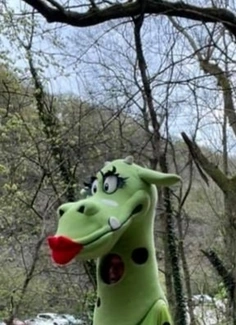 Person dressed as a green dragon with red lips, standing outdoors with trees in the background